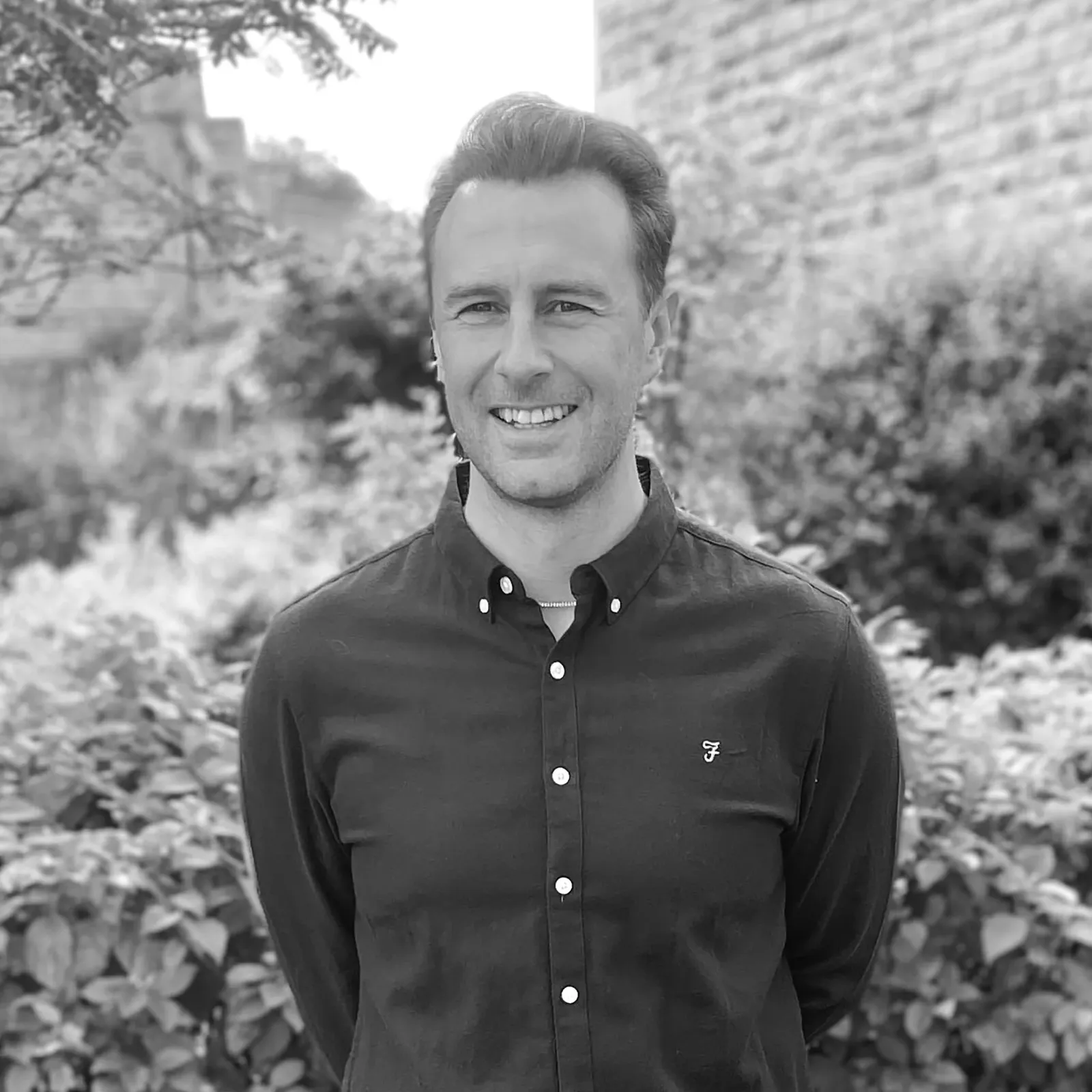 Man smiling outdoors, wearing a dark shirt, with a blurred garden background. Black and white photo.