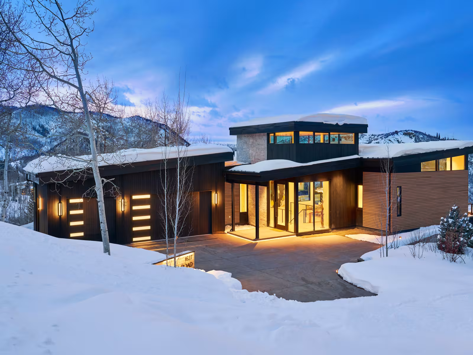 Modern house with glowing windows, surrounded by snowy landscape at dusk.