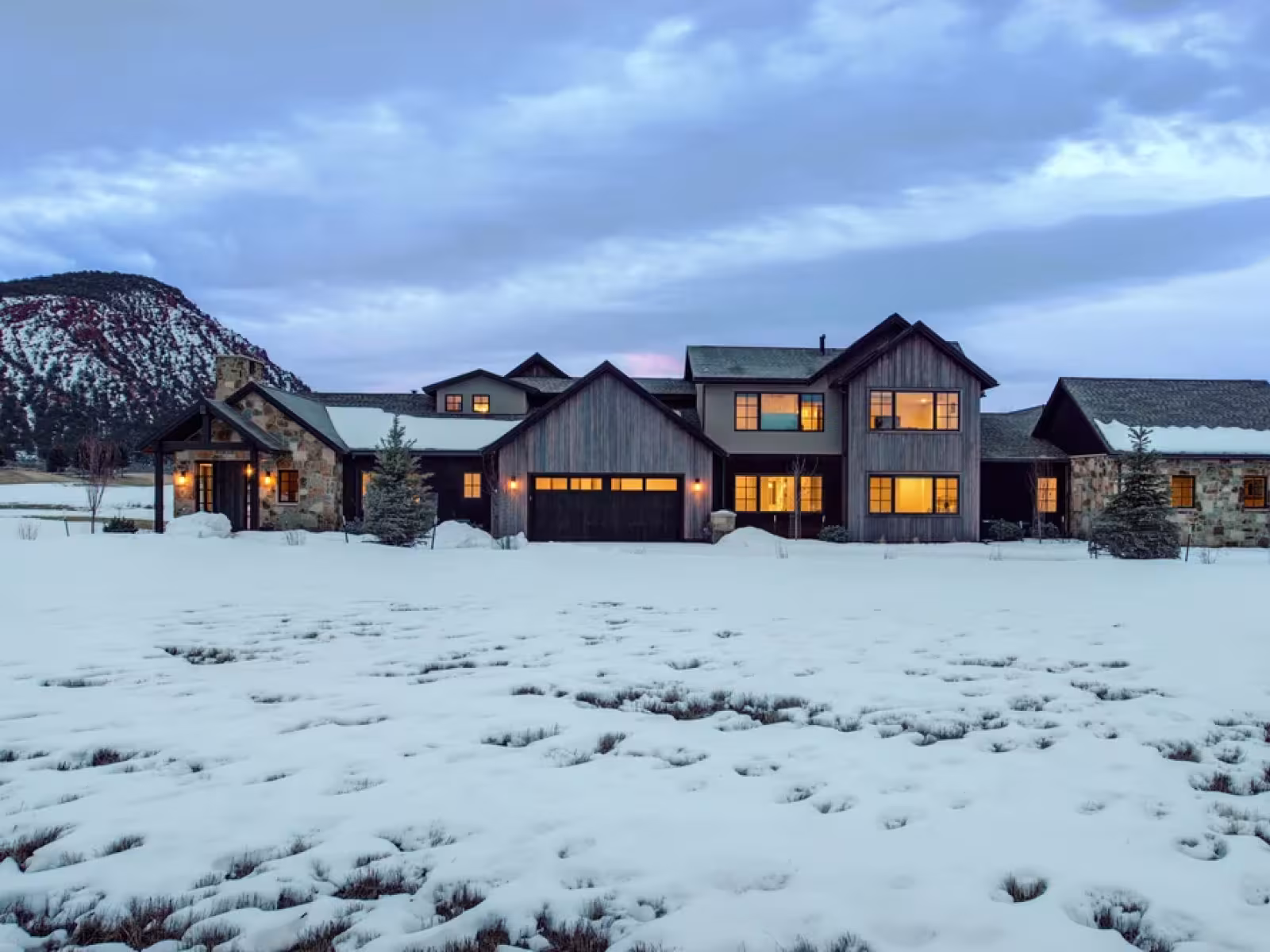 Snow-covered landscape with a warmly lit country house at dusk.