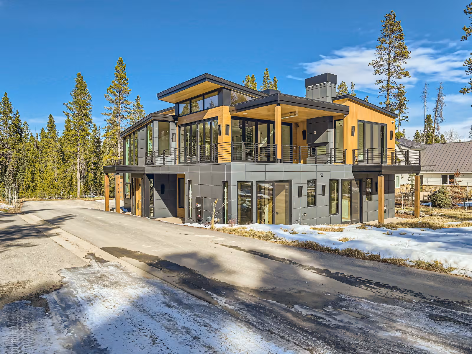 Modern house with large windows, surrounded by trees and light snow, under a clear blue sky.