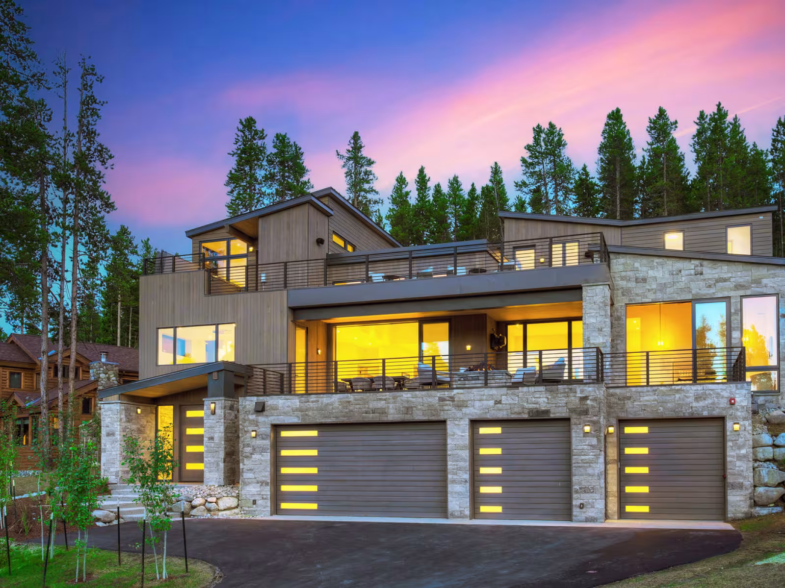 Modern house with large windows, illuminated at dusk, surrounded by tall trees.