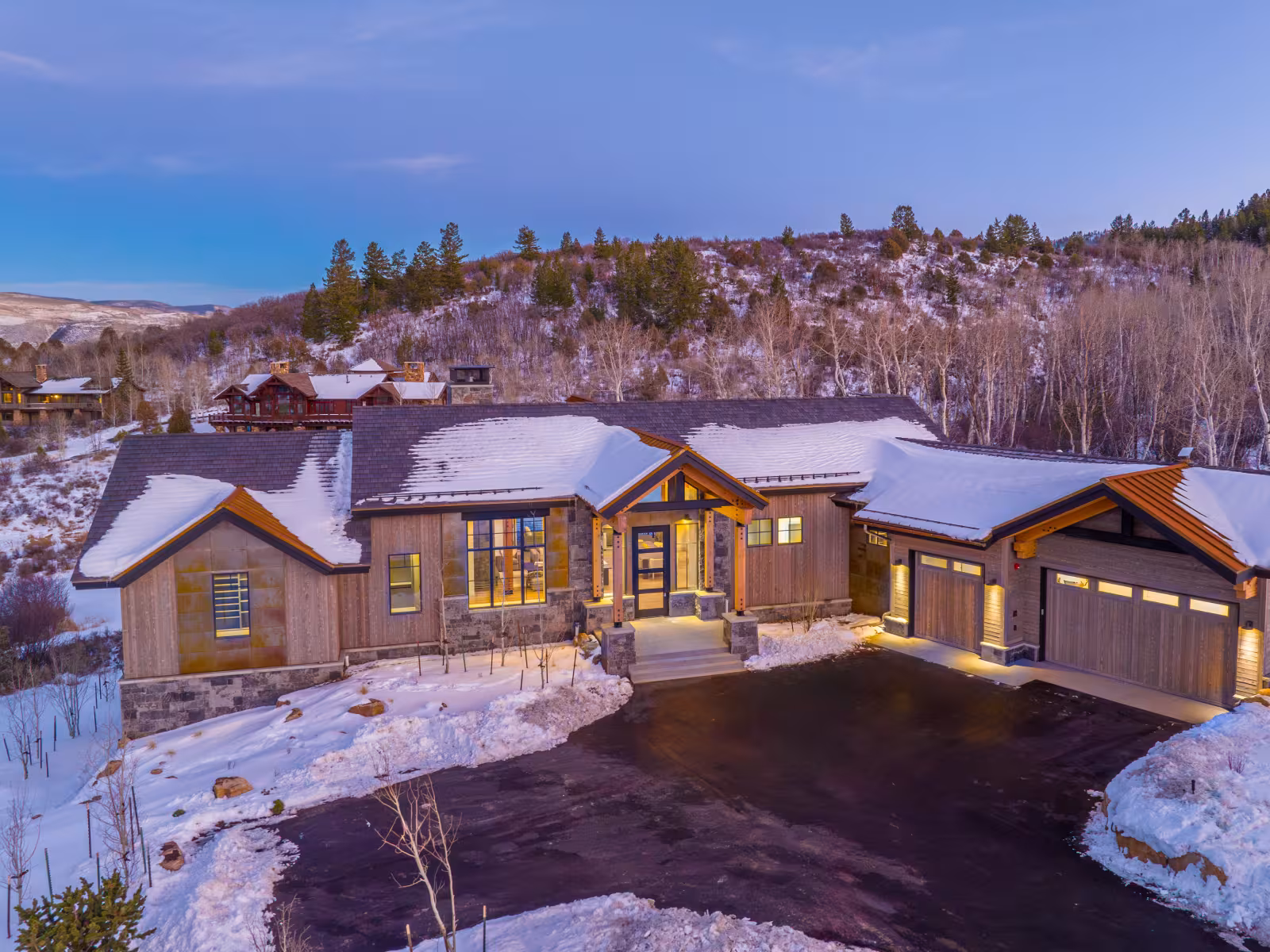 Snowy cabin at dusk with warm lit windows, surrounded by trees and hills.