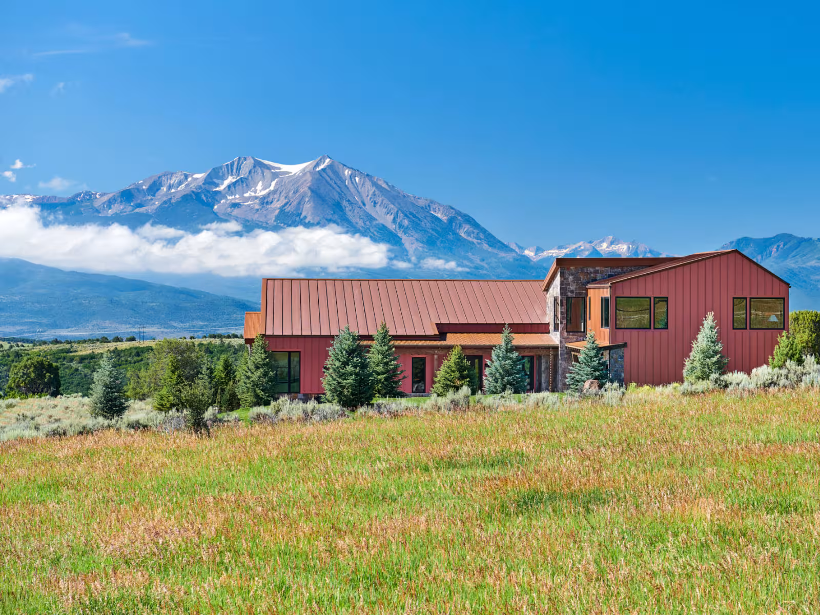 Modern house with mountains in background, sunny day, green field in foreground.