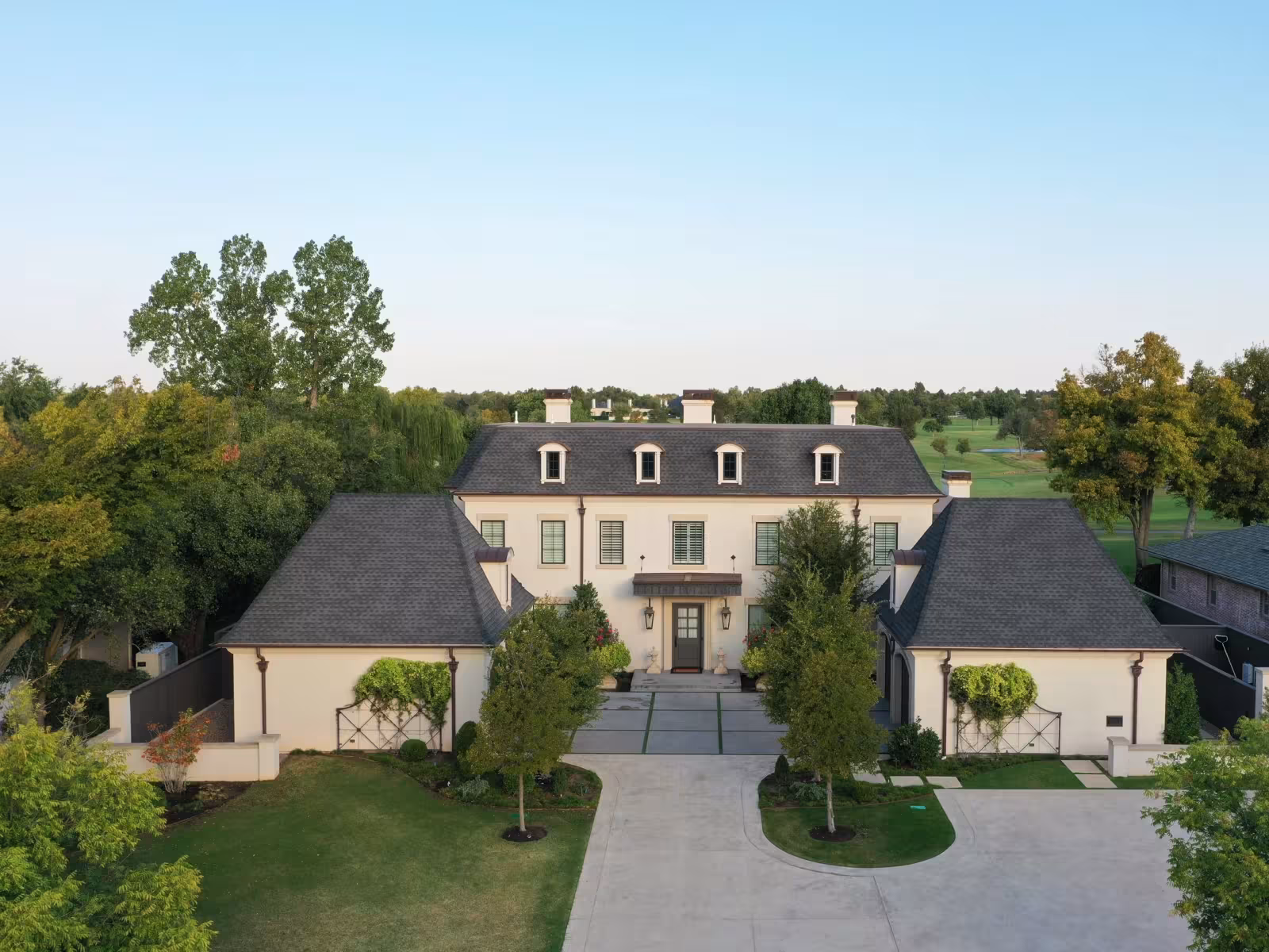 Large white house with dark roof, driveway, and surrounding trees.