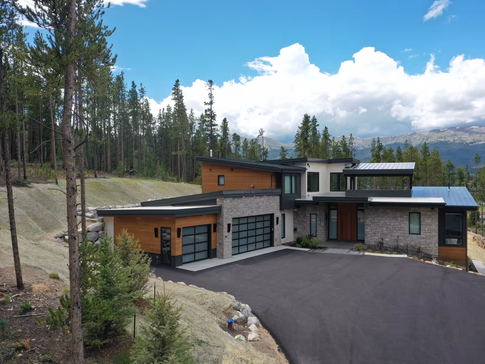 Modern house with large windows, surrounded by trees and mountains under a cloudy sky.