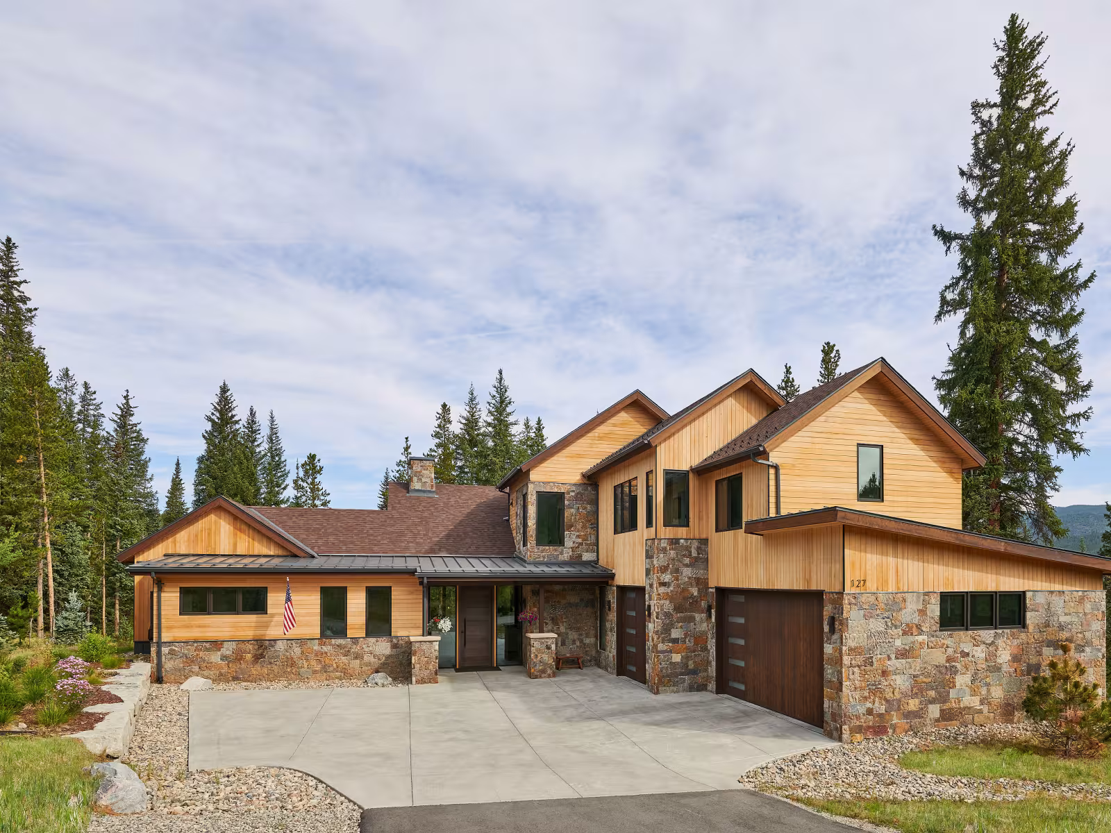 Two-storey wooden house with stone accents, surrounded by pine trees.