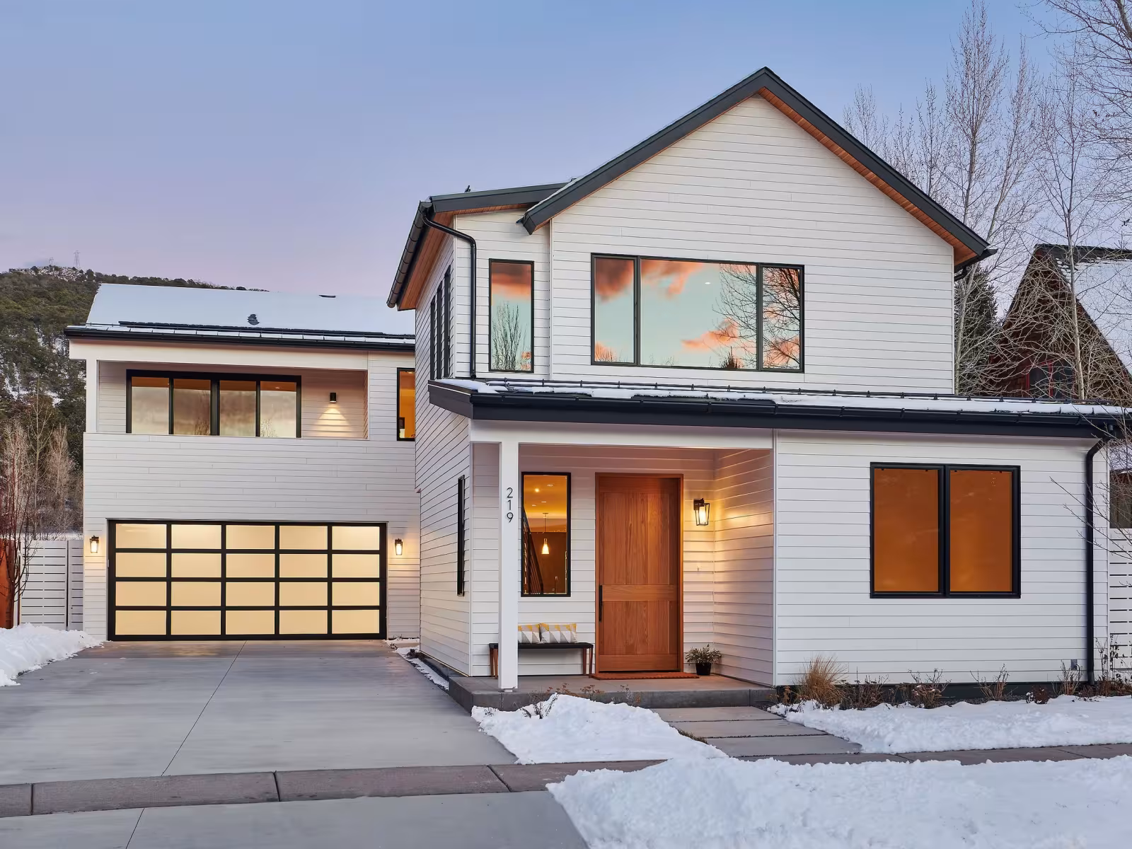 Modern white house with large windows, snowy driveway, and a double garage at dusk.