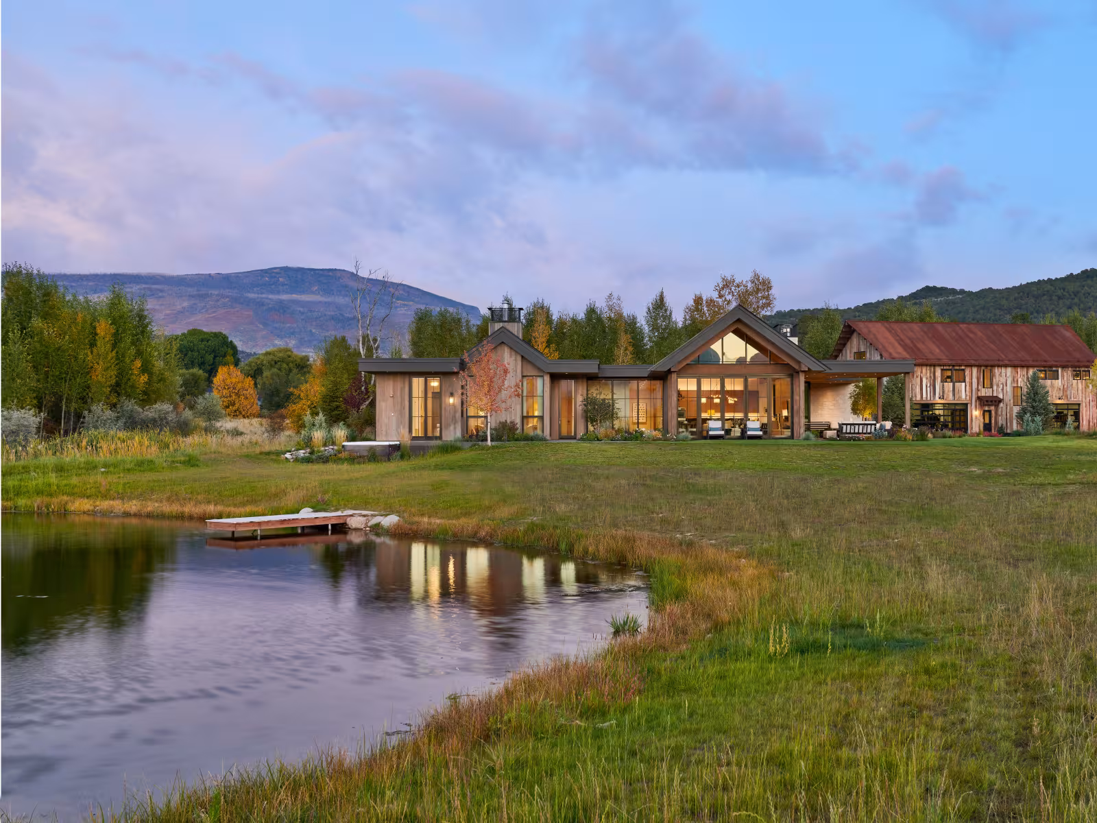 Rural house at sunset beside a pond, with mountains in the distance.