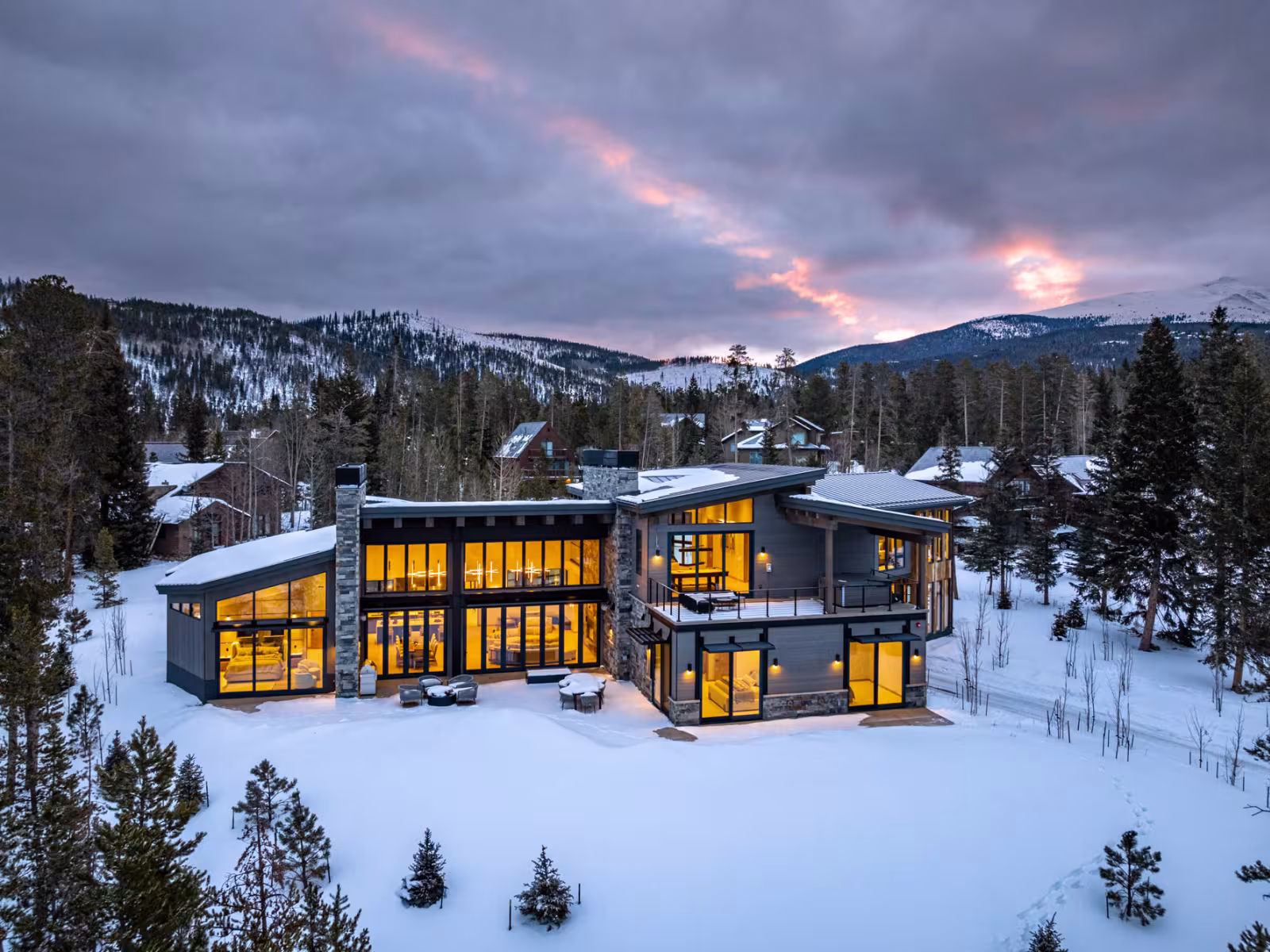 Modern house with lit windows, surrounded by snow and trees, under a cloudy sunset sky.