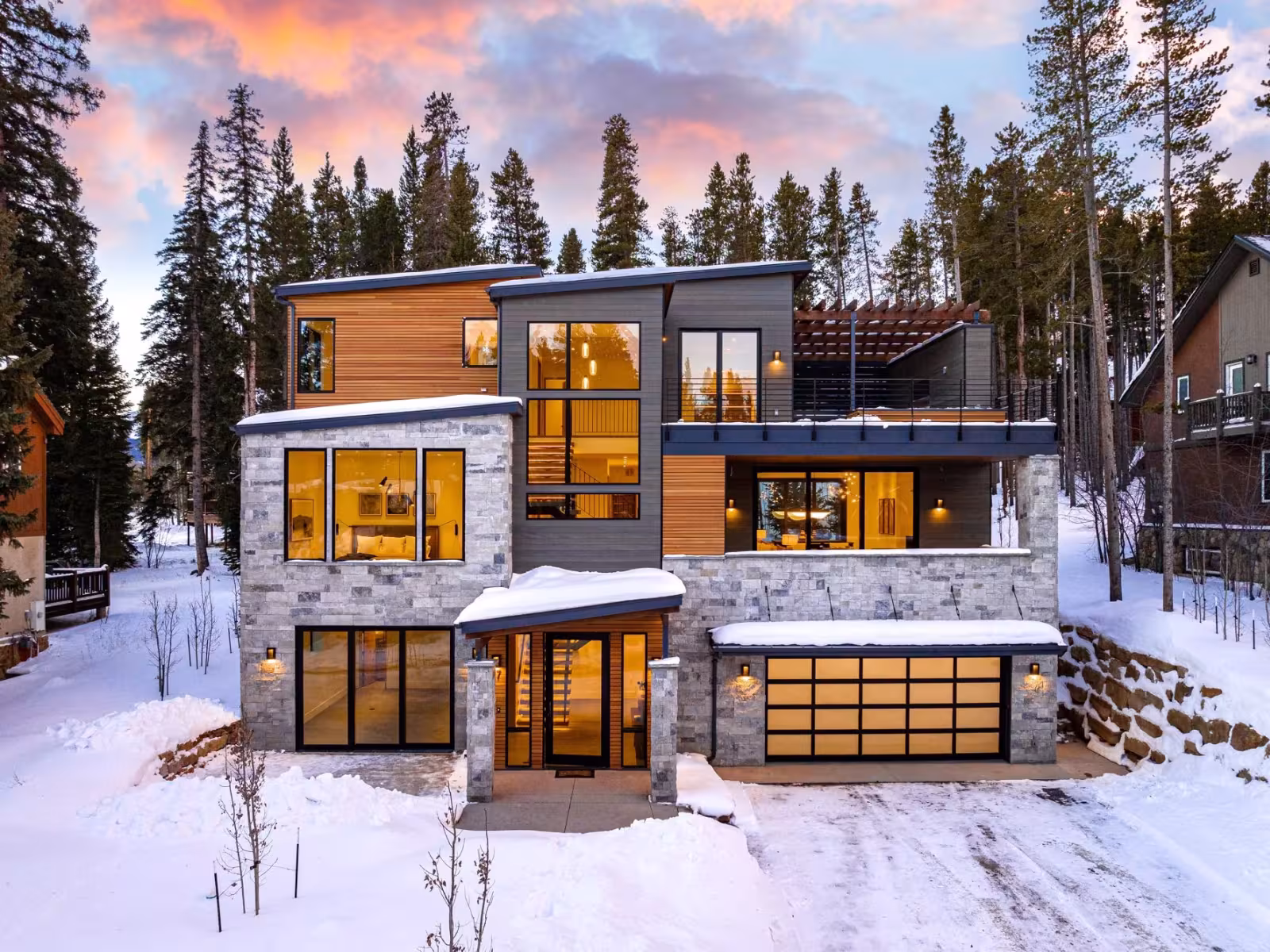 Modern house in snowy forest at sunset, warm lights glowing inside.