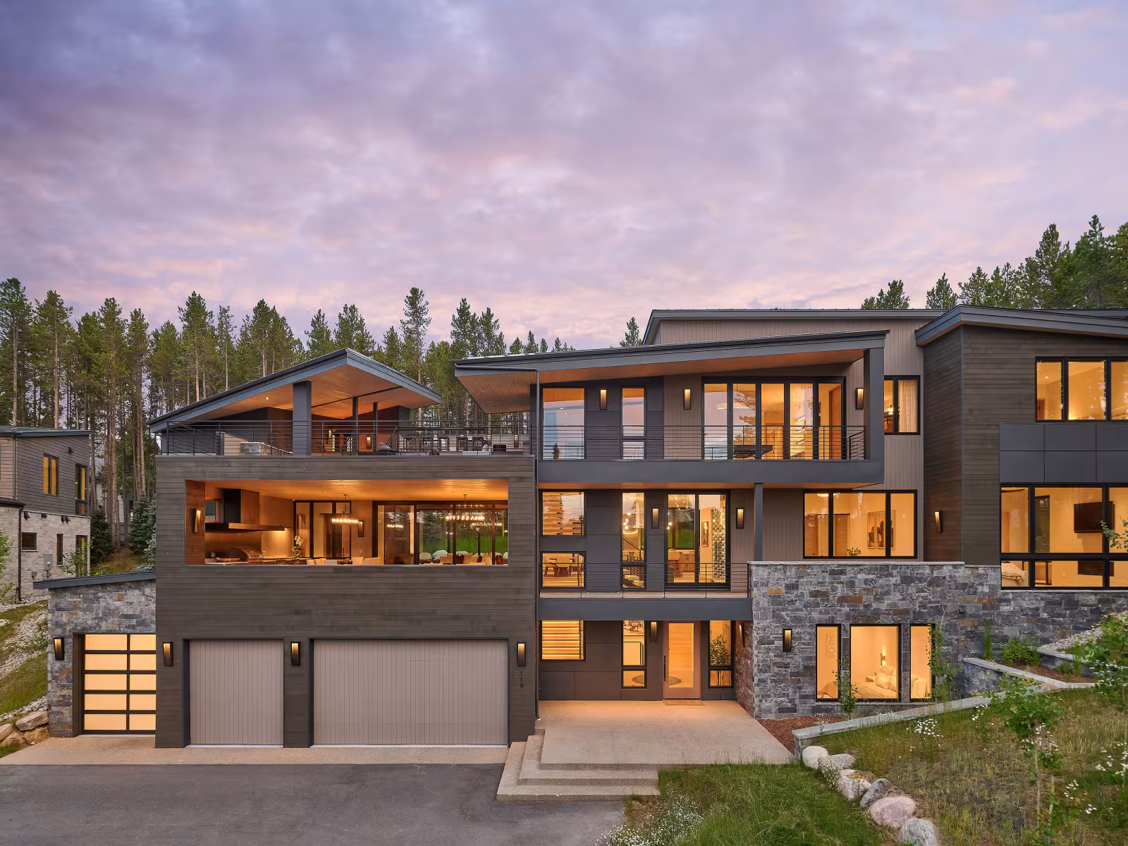 Modern three-storey house, large windows, surrounded by trees, dusk sky.