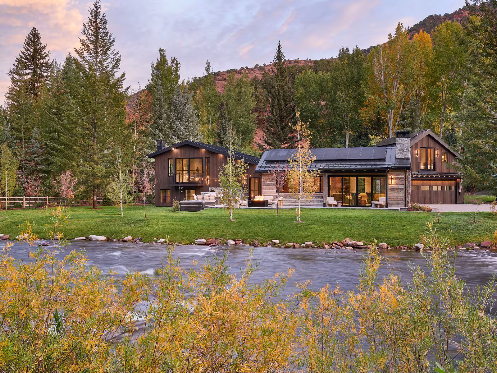 Modern riverside cabin surrounded by trees under a colourful sunset.