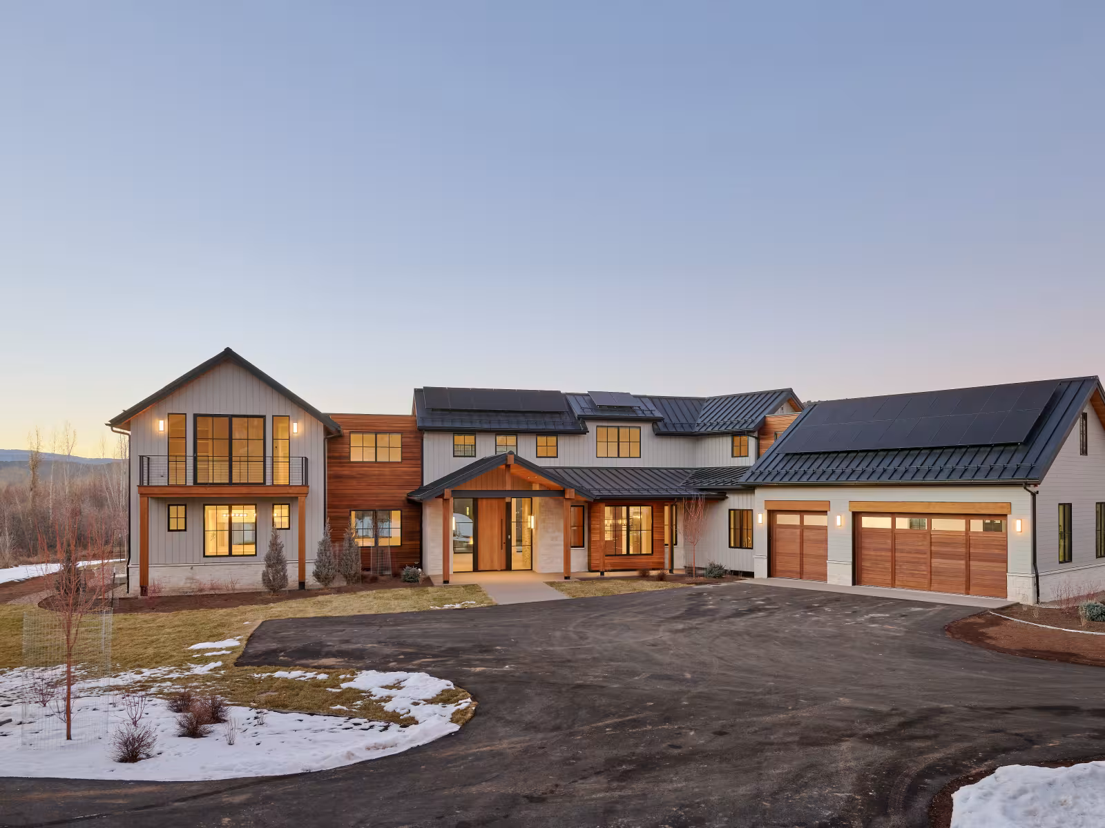 Modern house with large windows, solar panels, and snowy landscape at dusk.