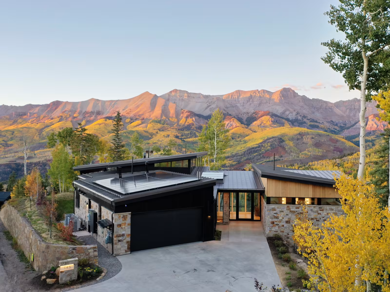 Modern house with mountain backdrop at sunset, autumn trees surrounding.